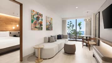 A modern hotel room with a bed, sofa, ottoman, and a dining area with a table and chairs. There are two large paintings on the wall and a TV mounted on the wall.