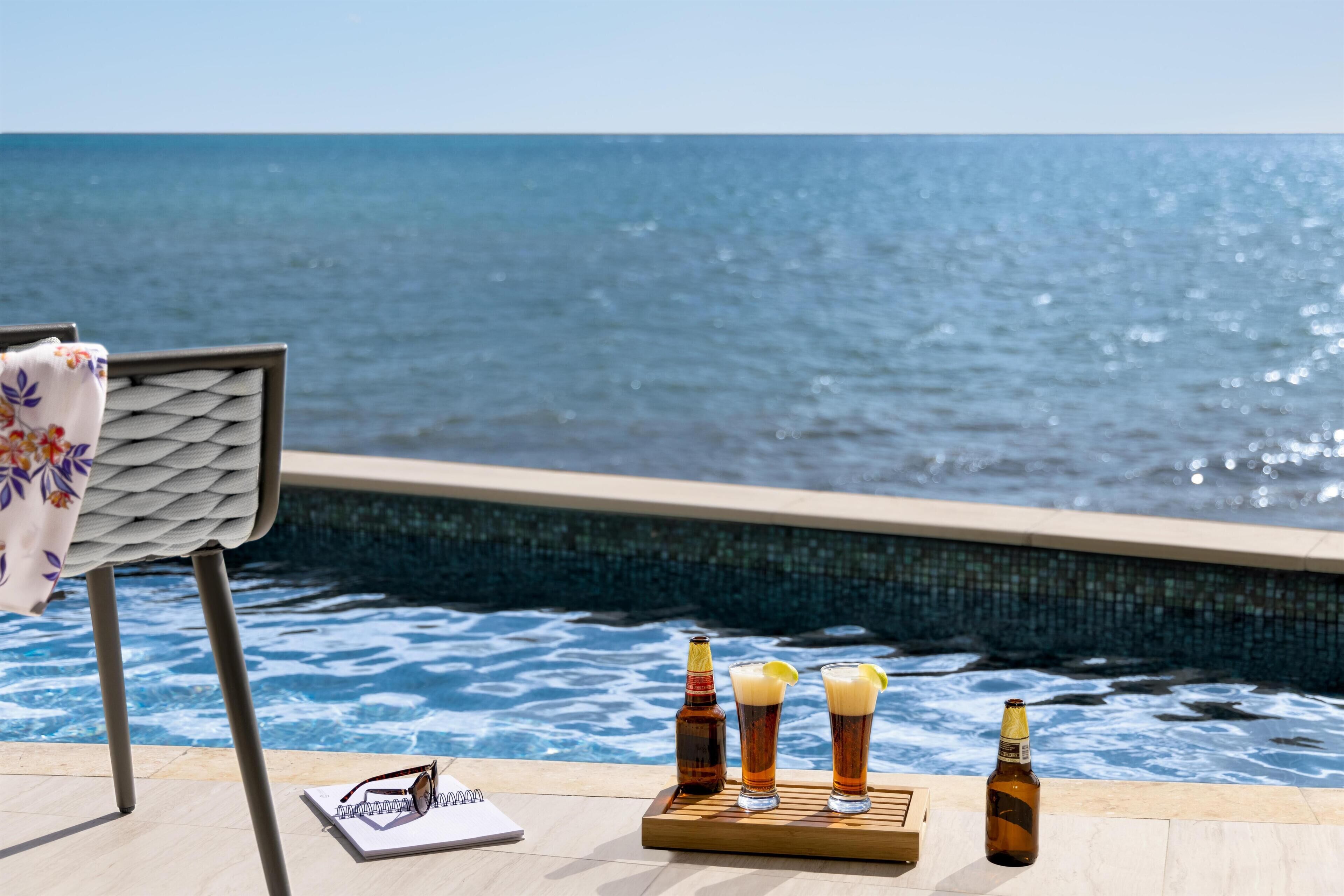 A poolside setting with a chair, a notebook, and three glasses of beer and a bottle on a tray.