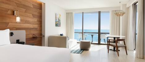 Habitación de hotel con una cama grande, un área de descanso con una mesa y sillas, un muro decorativo de madera y vistas al mar a través de amplios ventanales.