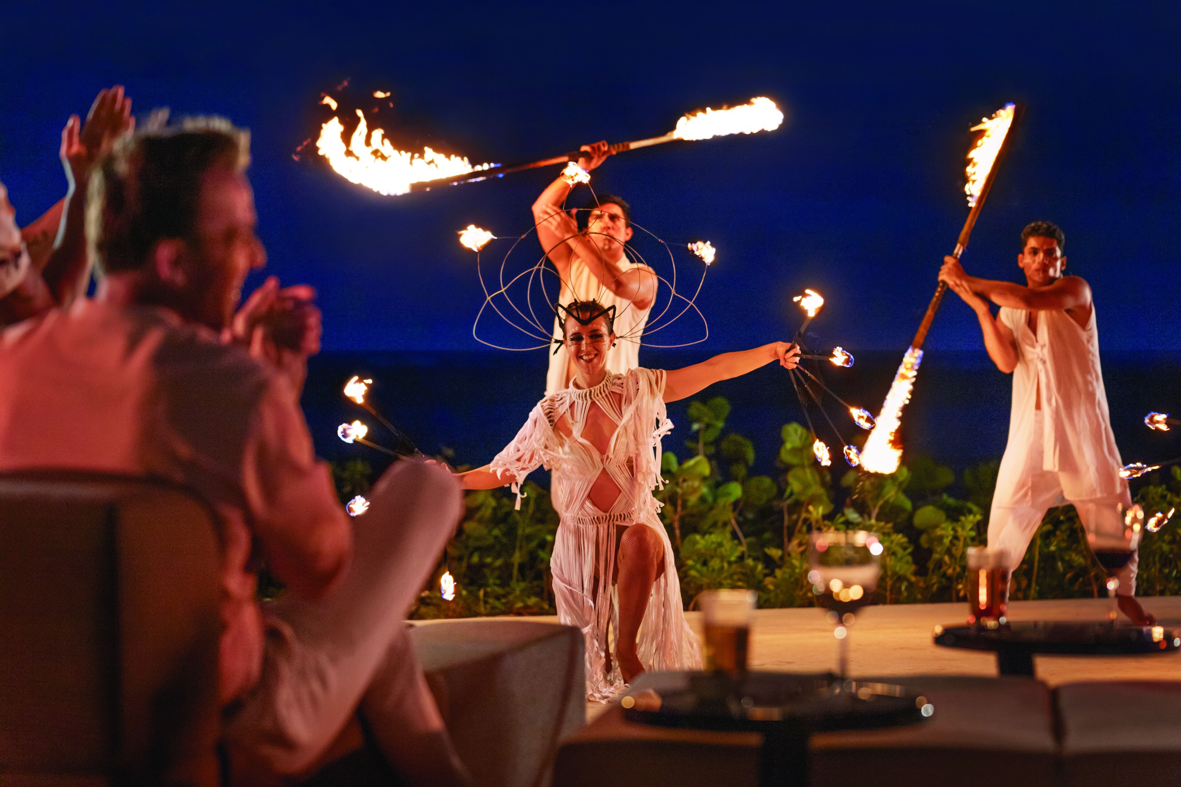 Un groupe de personnes fait une représentation en manipulant des torches enflammées, assis sur une terrasse aménagée avec des tables et des chaises.