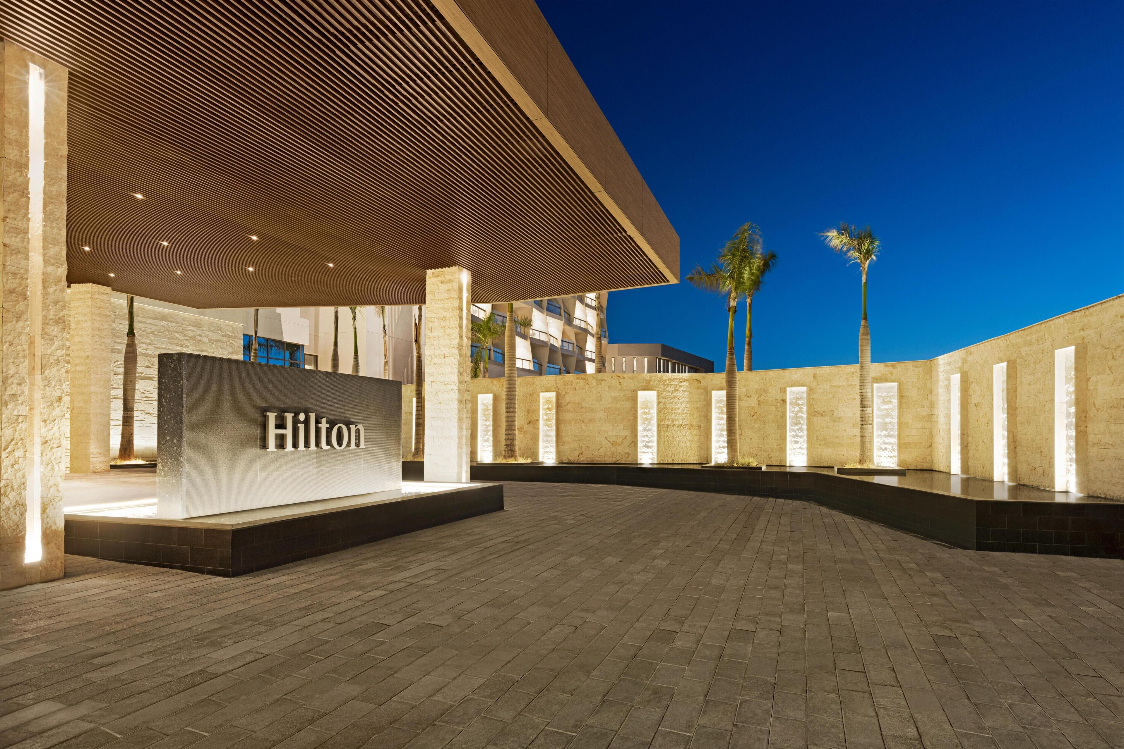 A modern hotel entrance with a prominent 'Hilton' sign, a brick-paved courtyard, and palm trees.