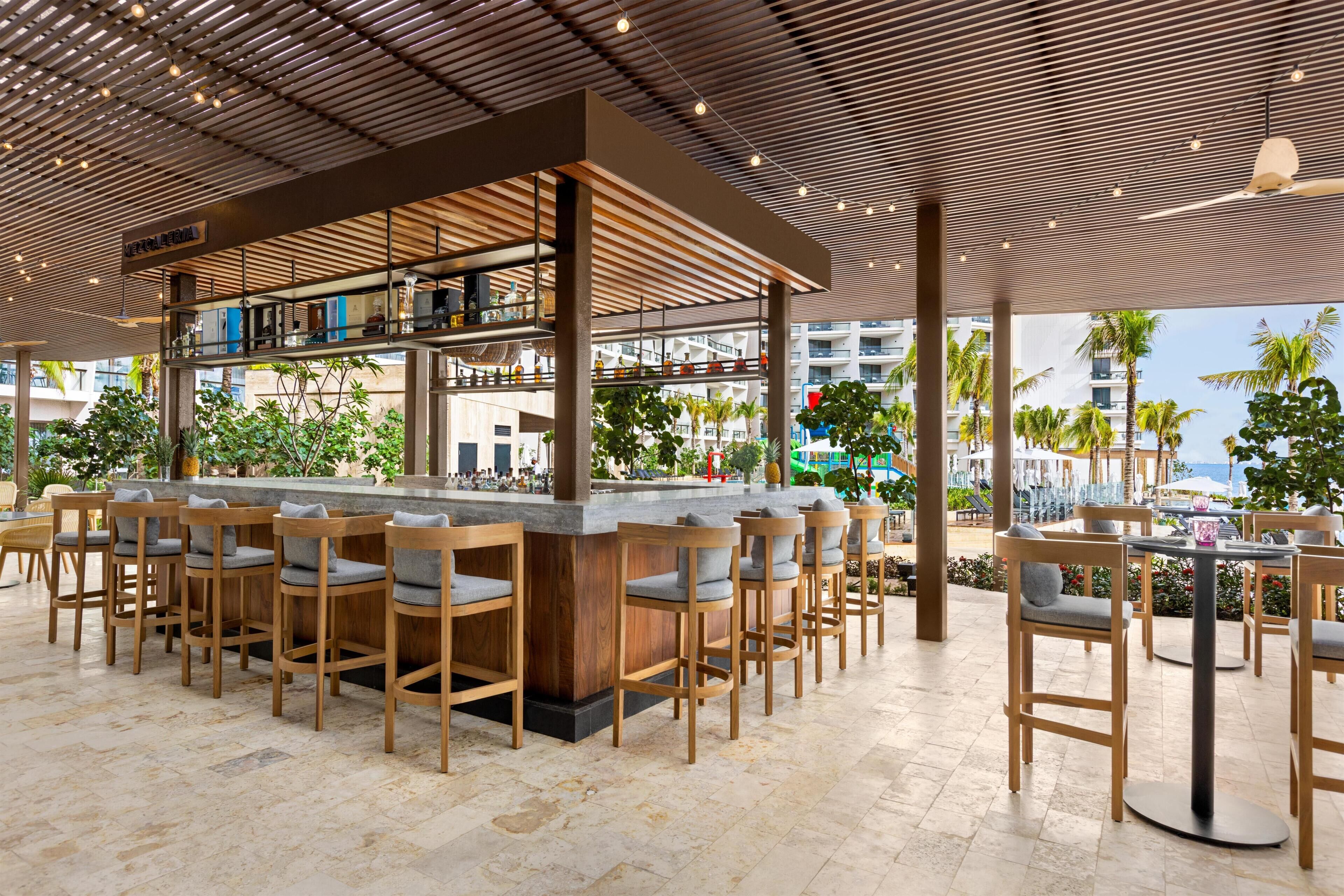 A bar area with wooden bar stools and a counter, surrounded by a covered outdoor seating area with tables and chairs.