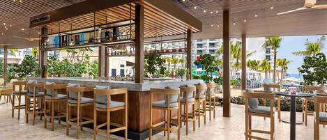 A bar area with wooden bar stools and a counter, surrounded by a covered outdoor seating area with tables and chairs.