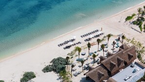 An aerial view of a beach resort with rows of sun loungers, palm trees, and thatched-roof structures.
