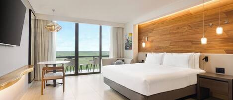 Una habitación de hotel moderna con una cama grande, un cabecero de madera, un rincón de comedor con mesa y sillas, y un balcón con vistas.
