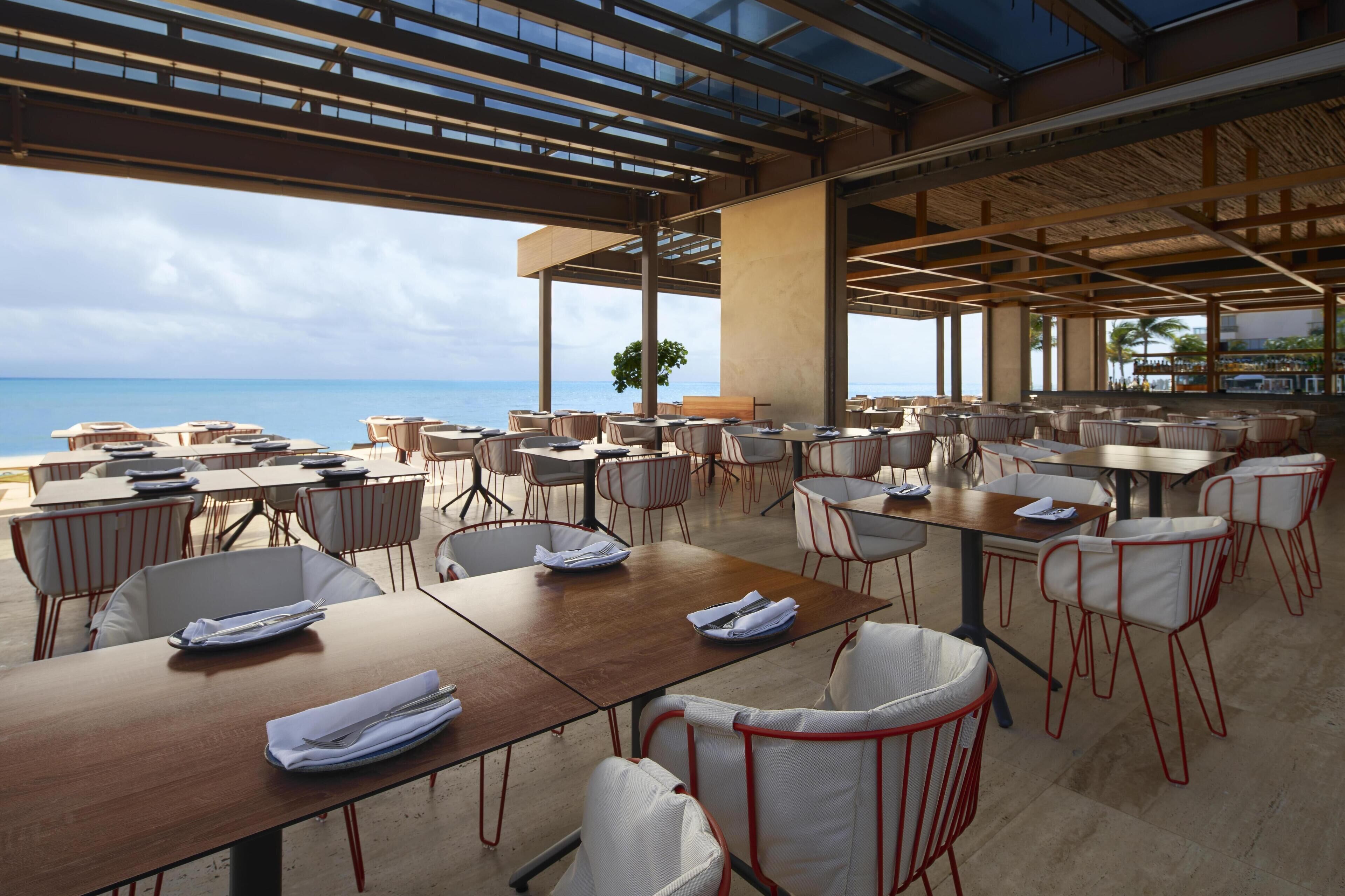 A spacious outdoor dining area with multiple tables and chairs, large windows offering a view of the ocean, and a modern architectural design with exposed beams and natural light.