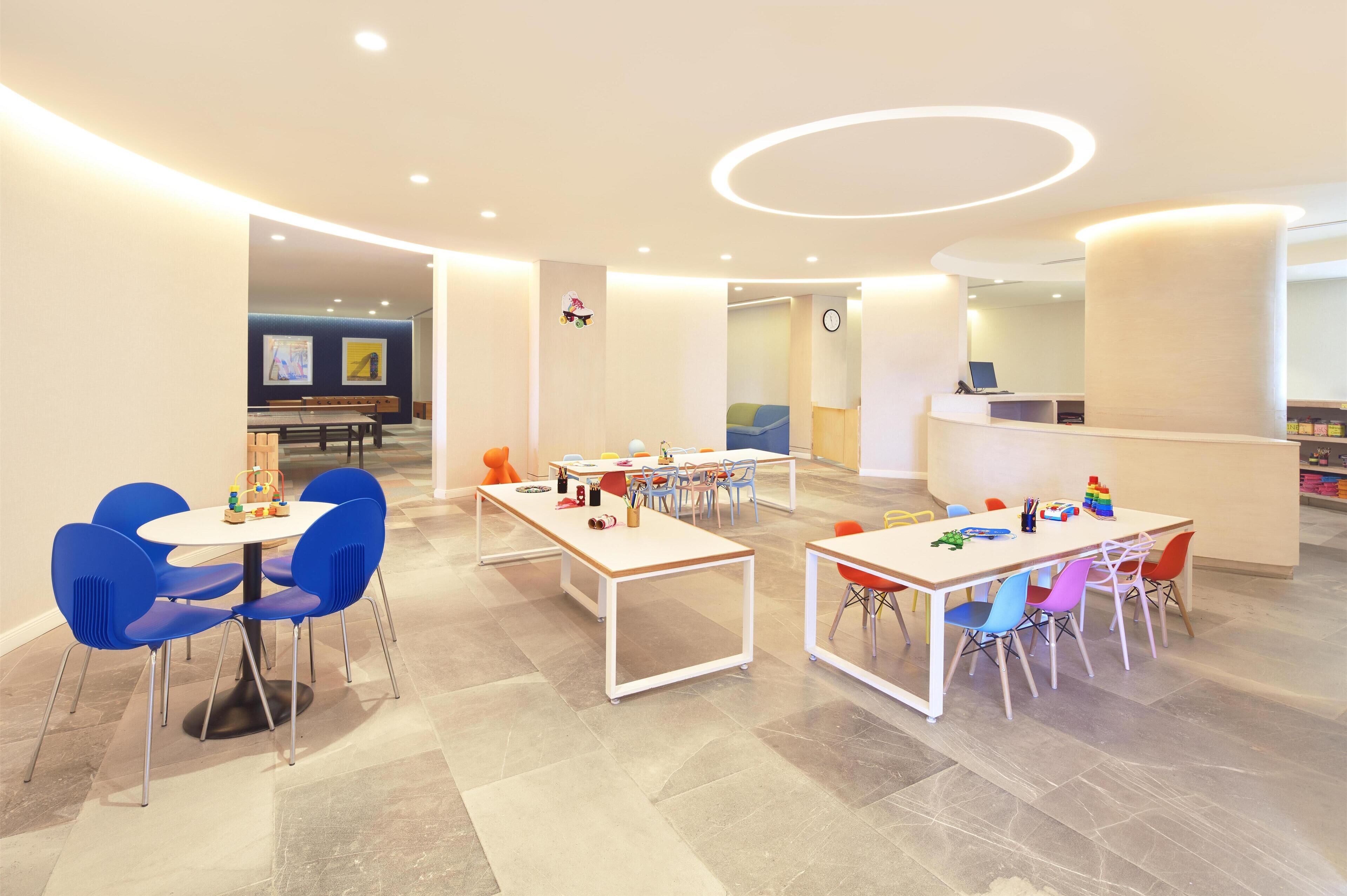 Un espace de jeux moderne pour enfants, équipé de tables et de chaises, de jouets et d’un comptoir d’accueil.