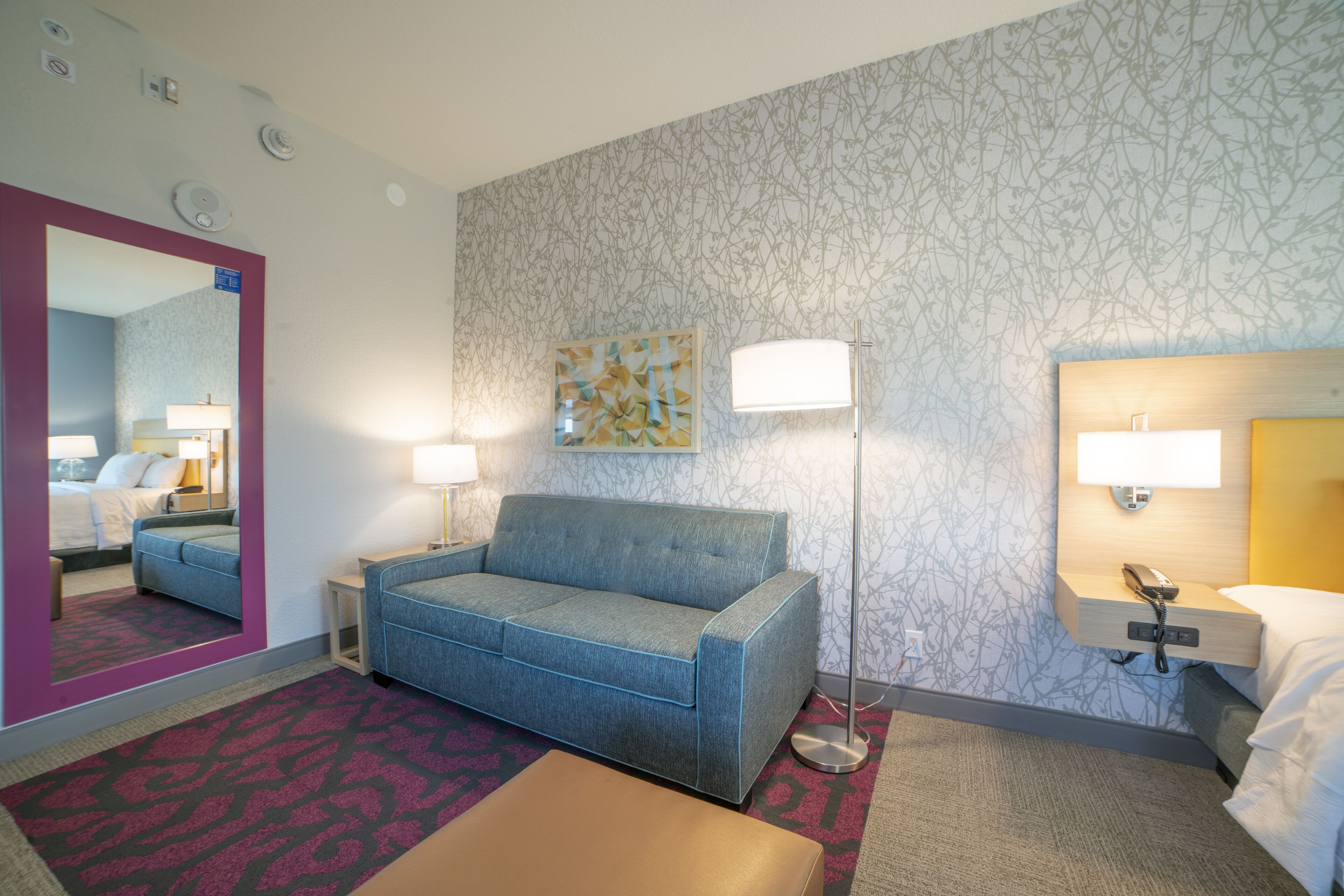 A hotel room with a blue sofa, a bedside table, a lamp, a mirror, and a patterned wall.