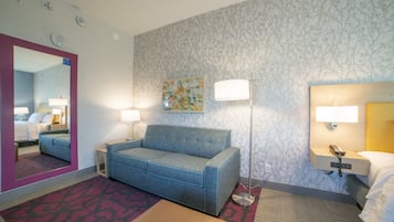 A hotel room with a blue sofa, a bedside table, a lamp, a mirror, and a patterned wall.