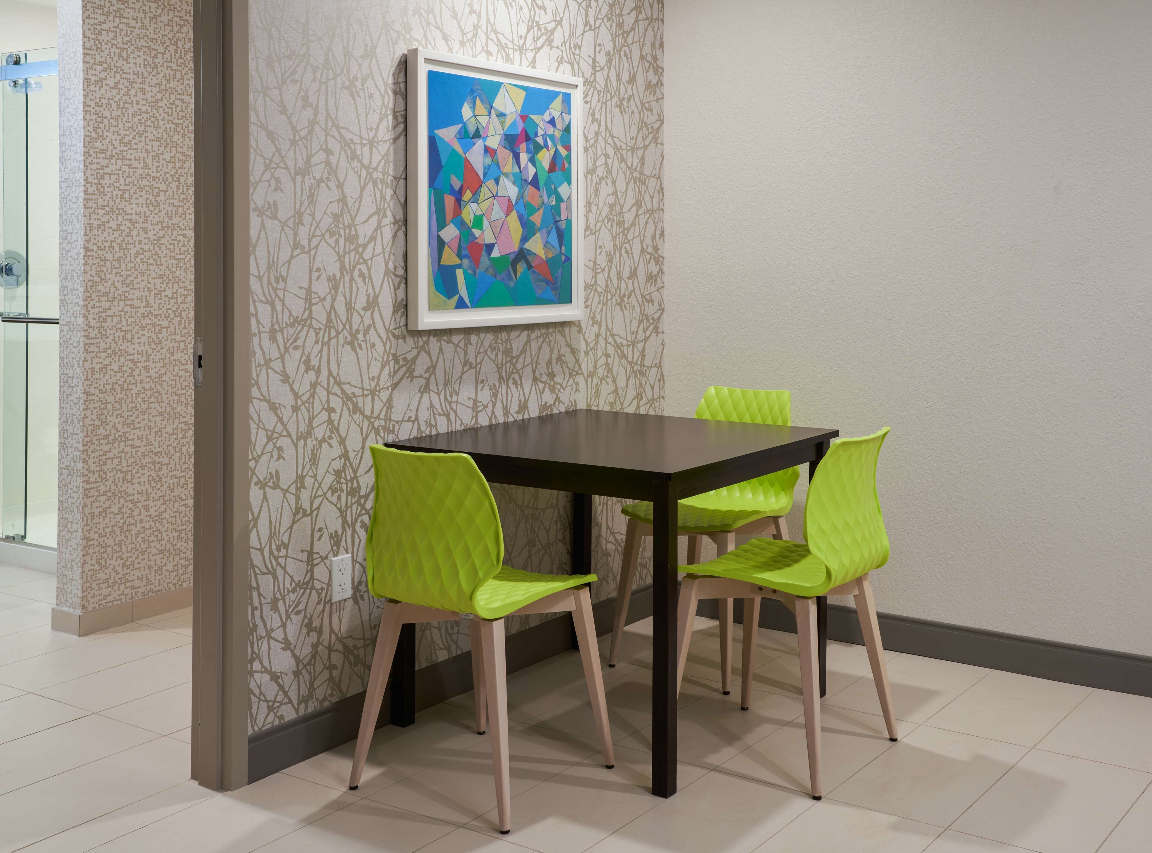 A dining area with a dark wooden table and four green chairs. A colorful abstract painting is on the wall above the table.