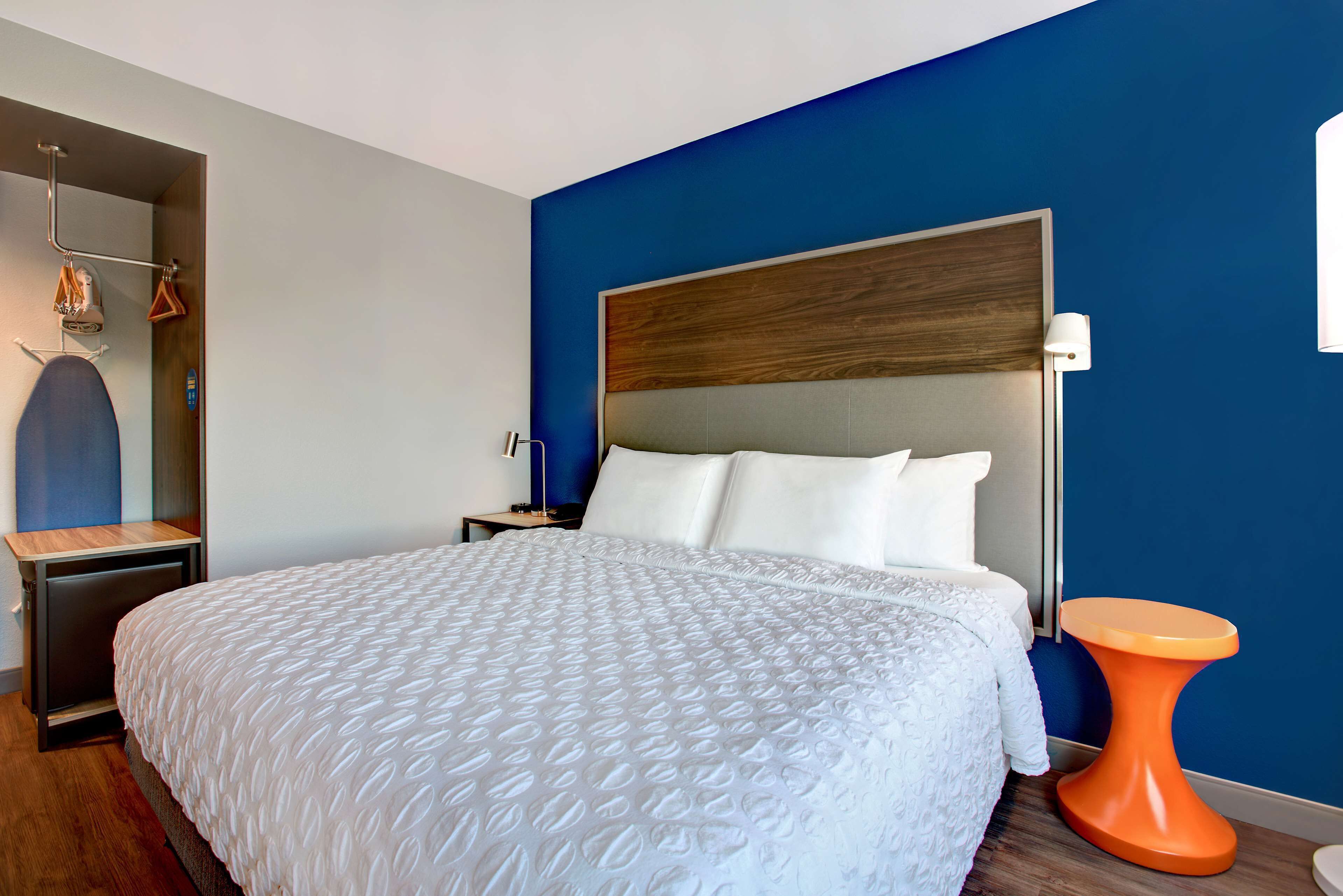 A modern hotel room with a large bed, a wooden headboard, a small orange side table, and a blue accent wall.
