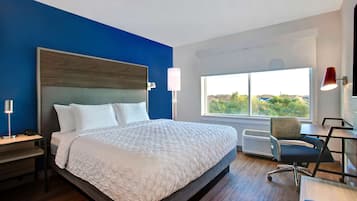 A modern hotel room with a large bed, a desk with a chair, a window with a view, and a red lamp.