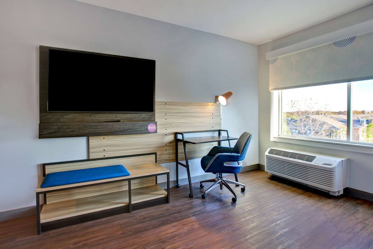 Un espacio de oficina moderno con un televisor de pantalla plana grande, un escritorio con silla, un banco y una ventana con vistas al exterior.