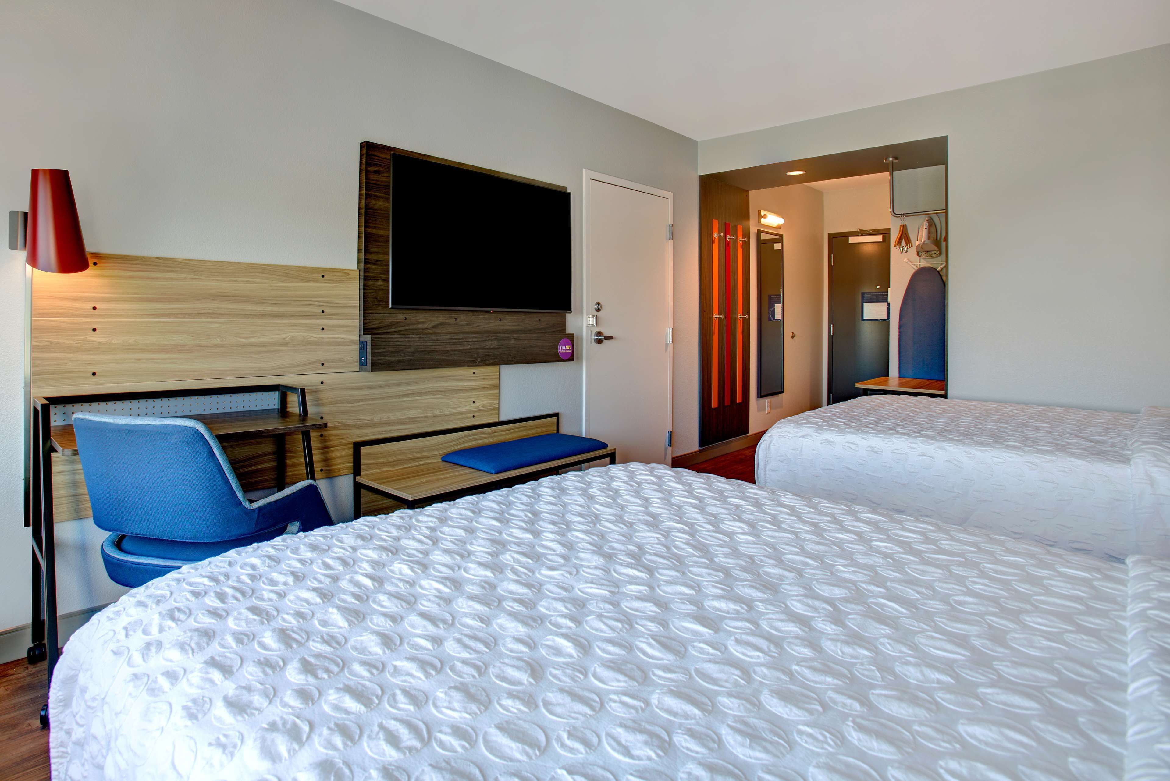 A hotel room with two beds, a desk, a chair, a TV mounted on the wall, and a door leading to another room.