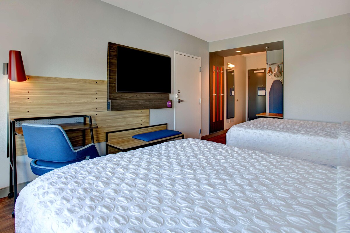 Habitación de hotel con dos camas, un escritorio, una silla, un televisor montado en la pared y una puerta que da a otra habitación.