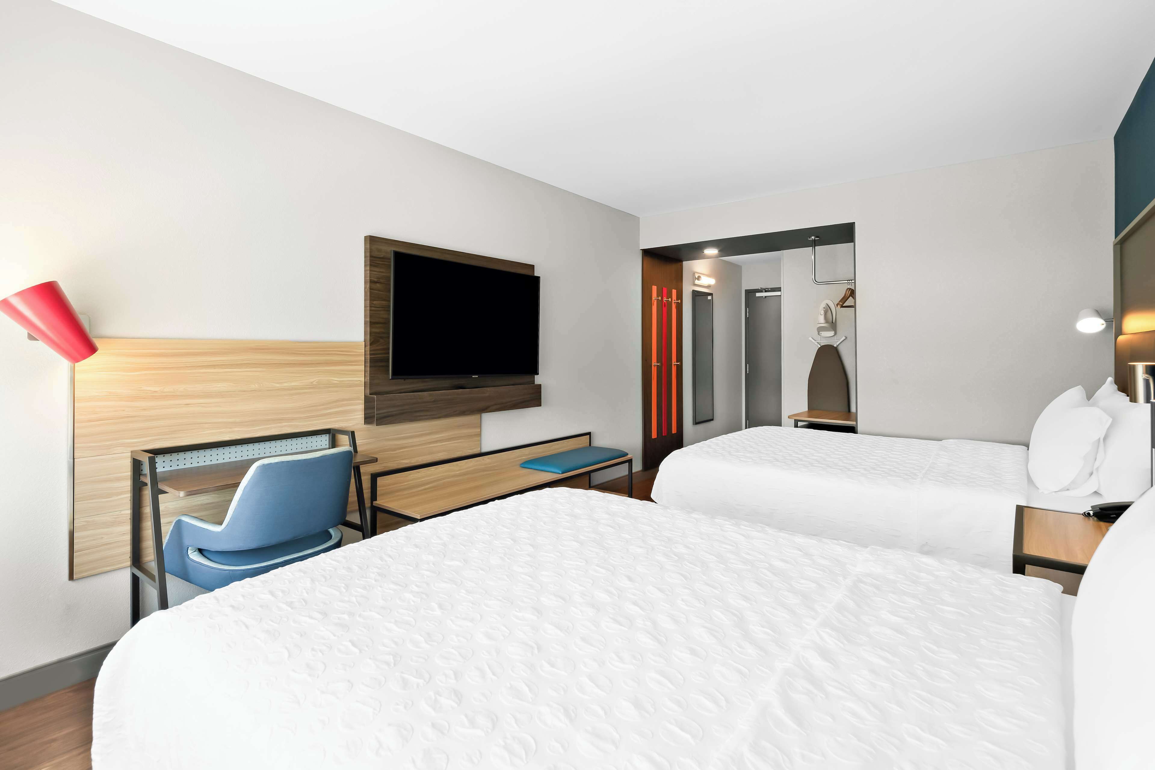 Habitación de hotel con dos camas, un televisor montado en la pared, un escritorio con una silla y un espejo.
