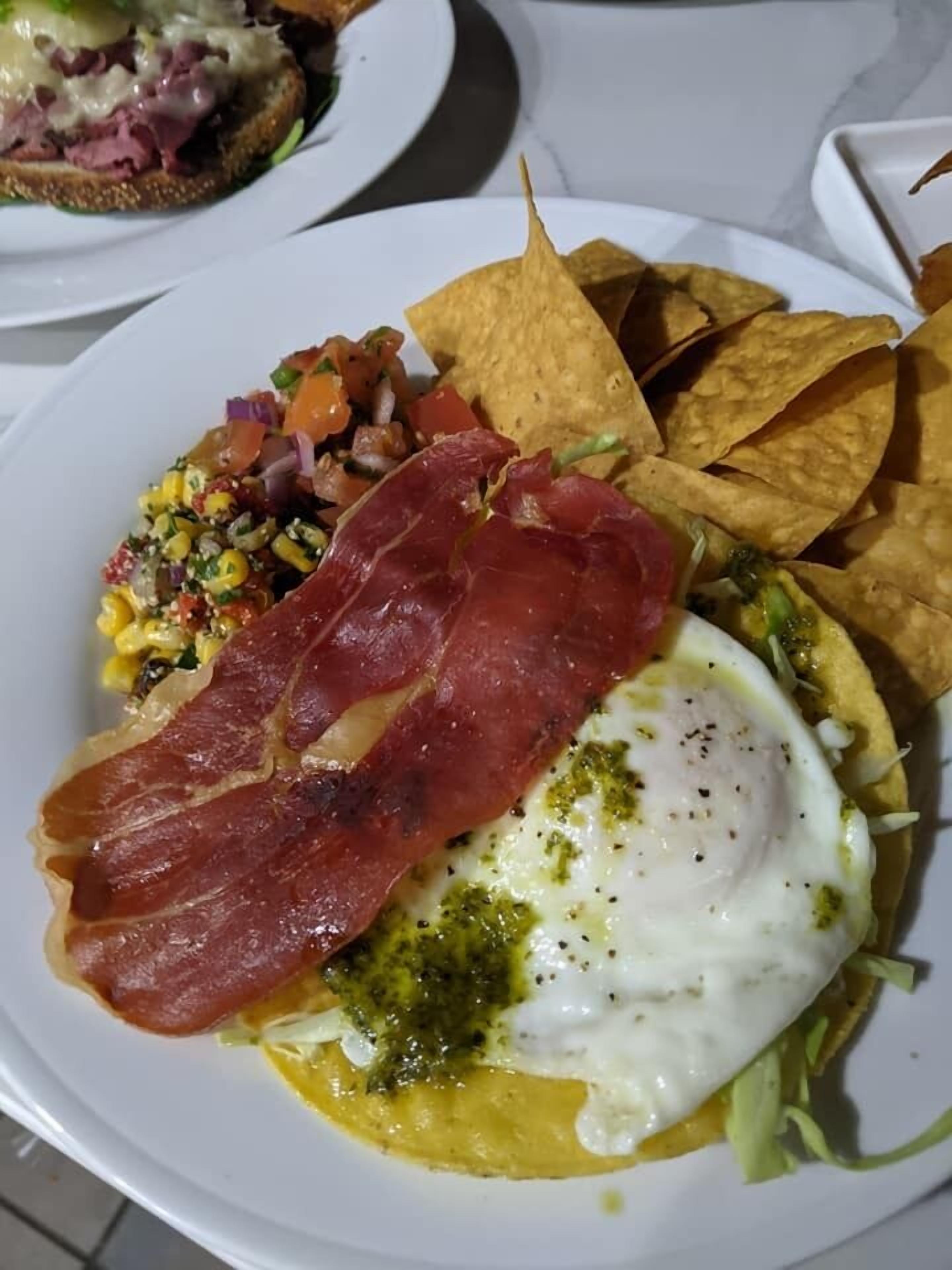 Une assiette avec un œuf au plat, une tranche de jambon de Parme, une salade de maïs et des chips de tortillas.