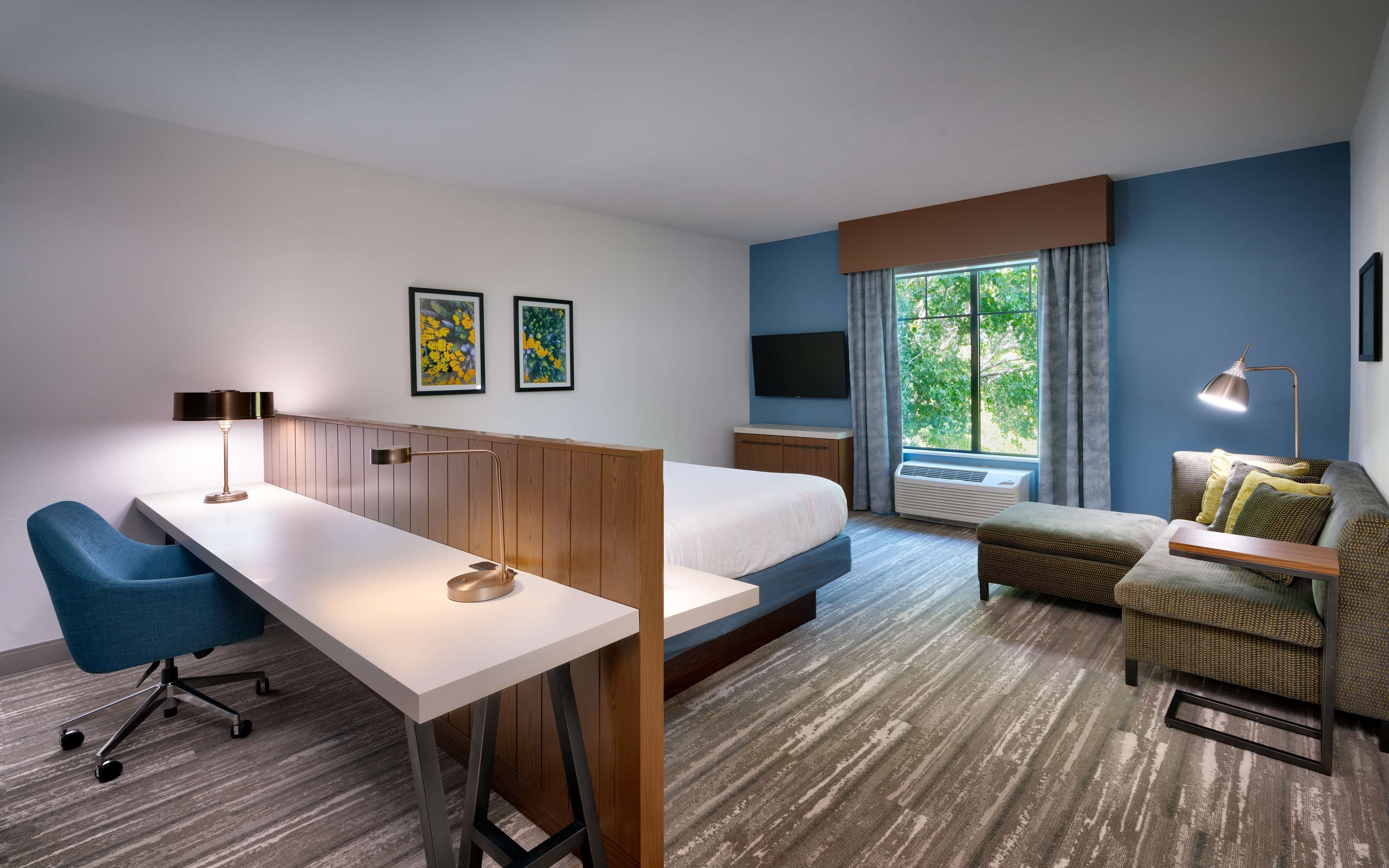 A hotel room with a bed, a desk, a chair, a TV, and a window with a view of trees.