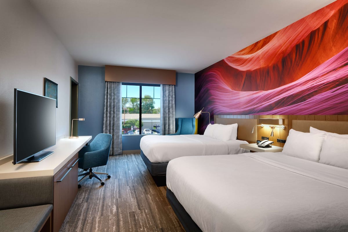 A hotel room with two beds, a desk with a TV, a chair, and a large window with curtains.