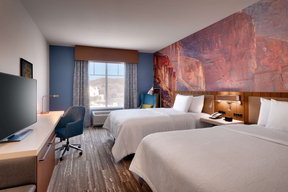 A hotel room with two beds, a desk with a flat-screen TV, a chair, a window with curtains, and a large abstract painting on the wall.