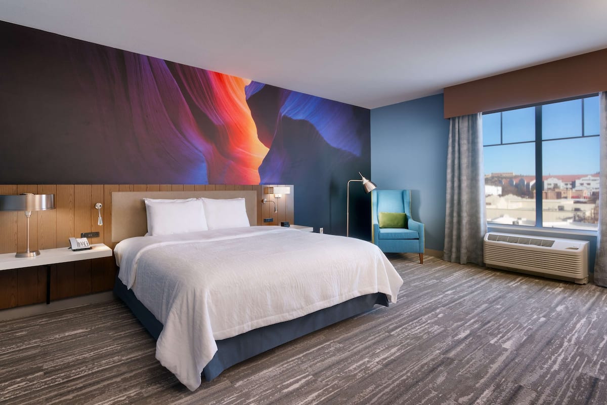 A modern hotel room with a large bed, a desk with a lamp, a chair, and a large window with a view.