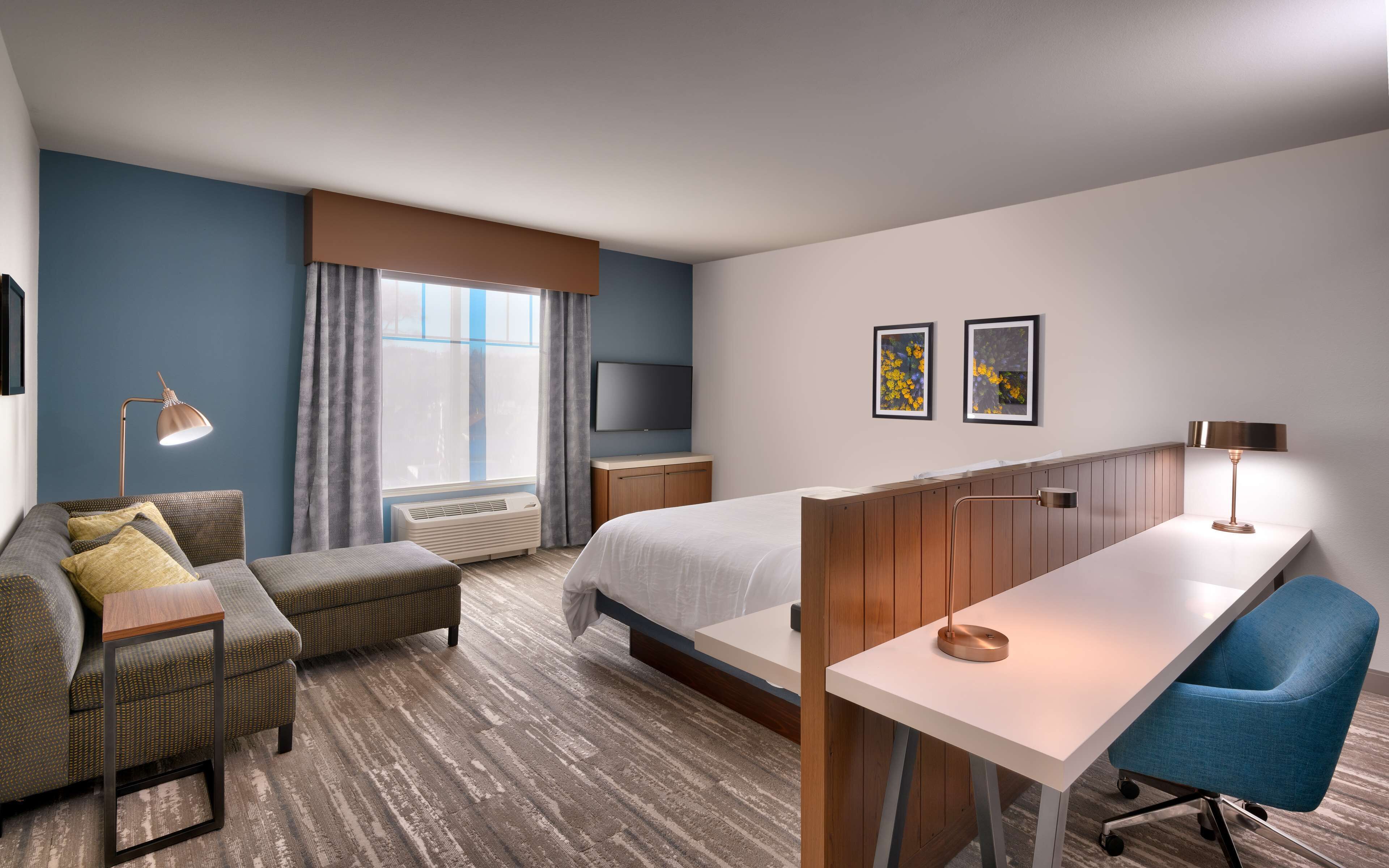 A hotel room with a bed, a desk, a chair, a sofa, a TV, and artwork on the wall.