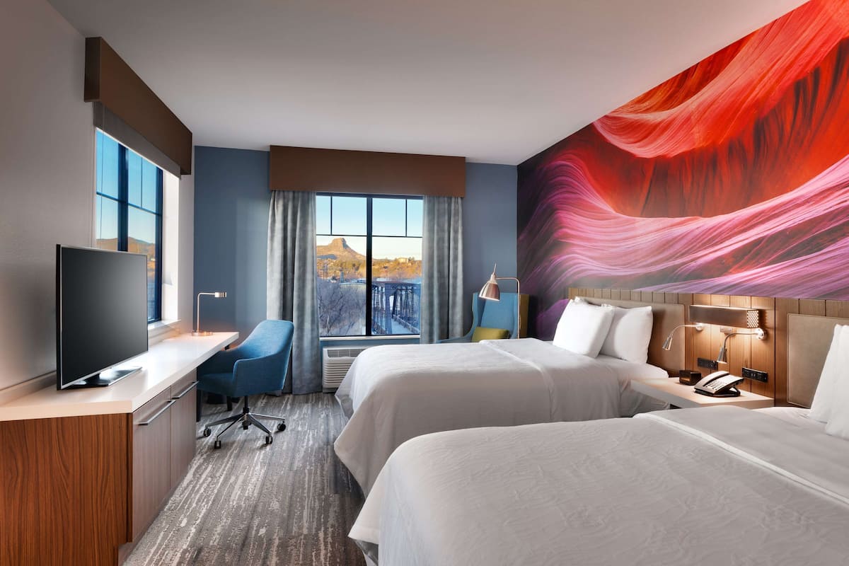 A hotel room with two beds, a desk, a chair, a TV, and a large window with a view of a bridge and mountains.