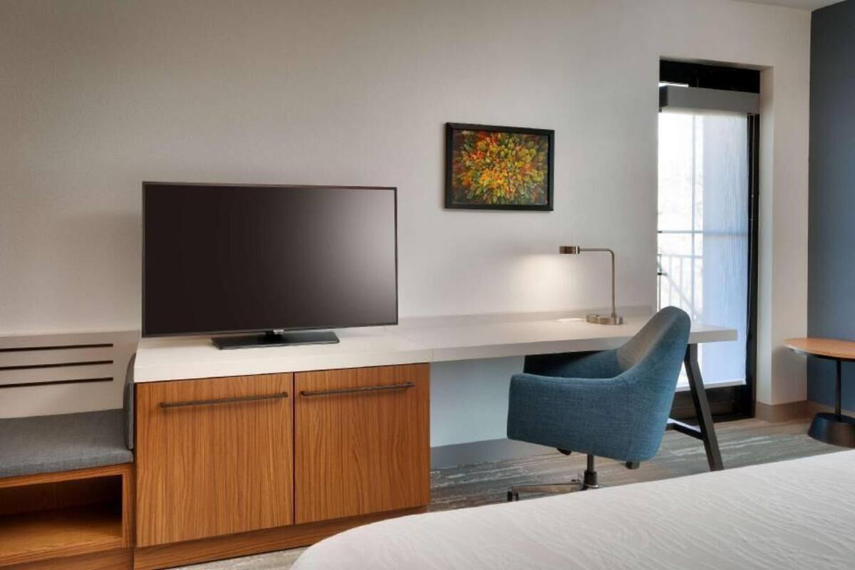 A modern hotel room with a flat-screen TV, a desk with a lamp, a blue chair, and a bed.