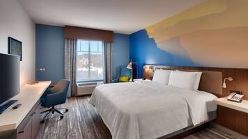 A hotel room with a bed, desk, chair, and a large window with a view of buildings.