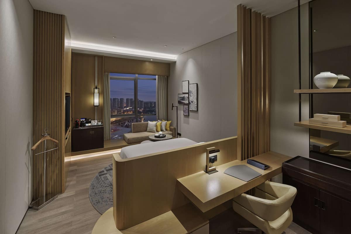 Une chambre d’hôtel moderne dotée d’un grand lit, d’un bureau, d’une chaise, d’une salle de bain et offrant une vue sur la ville au crépuscule.