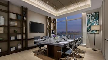 A modern conference room with a large table, chairs, a bookshelf, a flat-screen TV, and a view of the cityscape through large windows.
