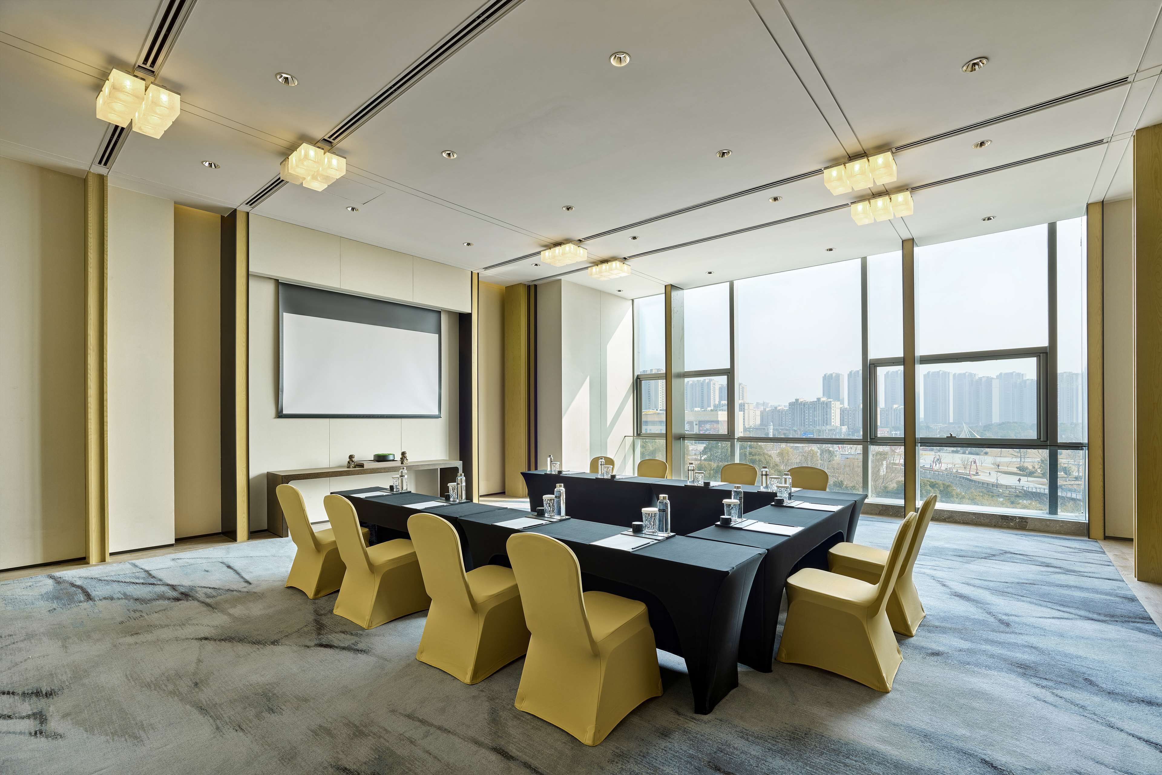 A conference room with a large table, yellow chairs, a projector screen, and large windows with a city view.