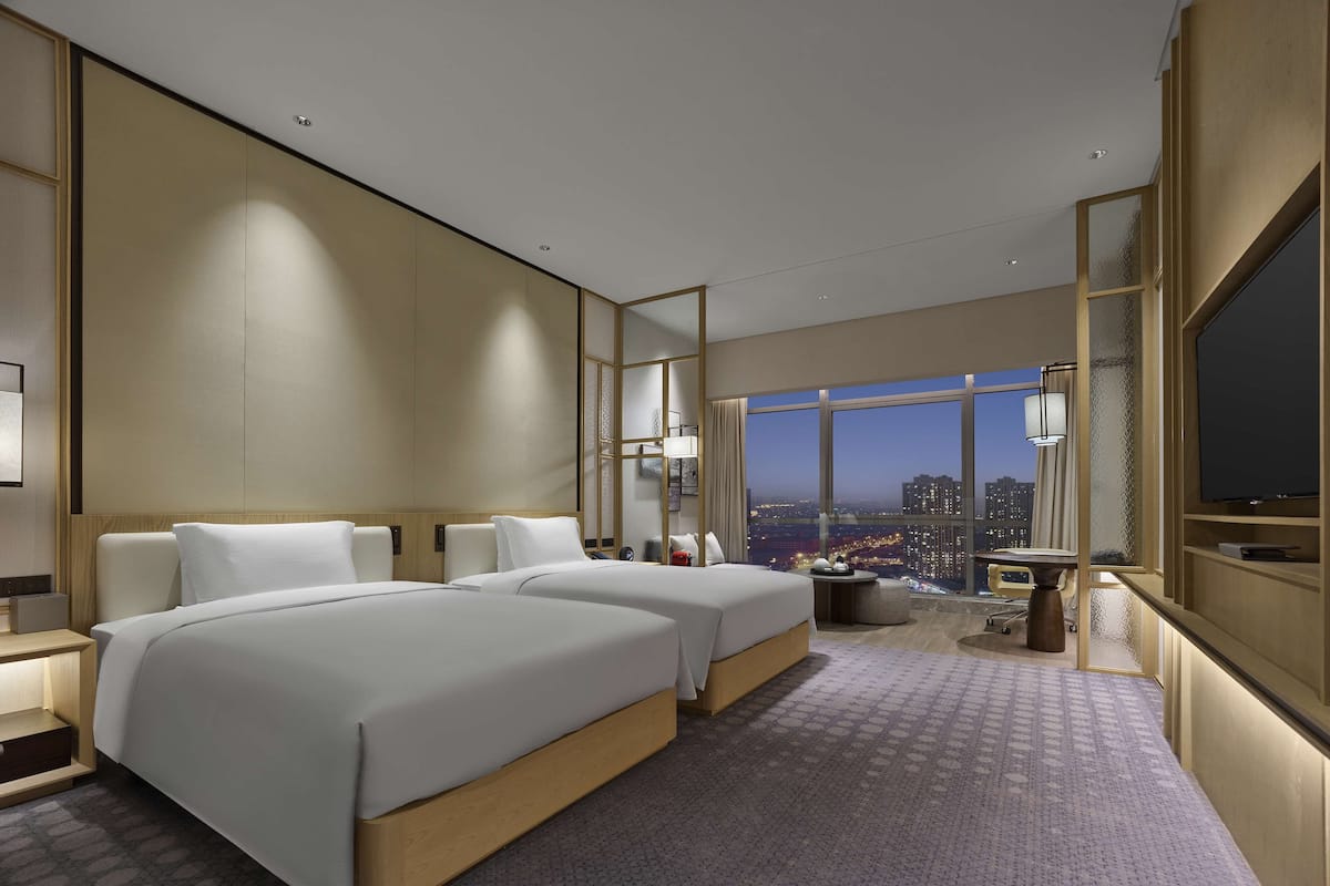 Une chambre d’hôtel moderne avec deux lits, une grande fenêtre donnant sur la ville, un bureau et une télévision.
