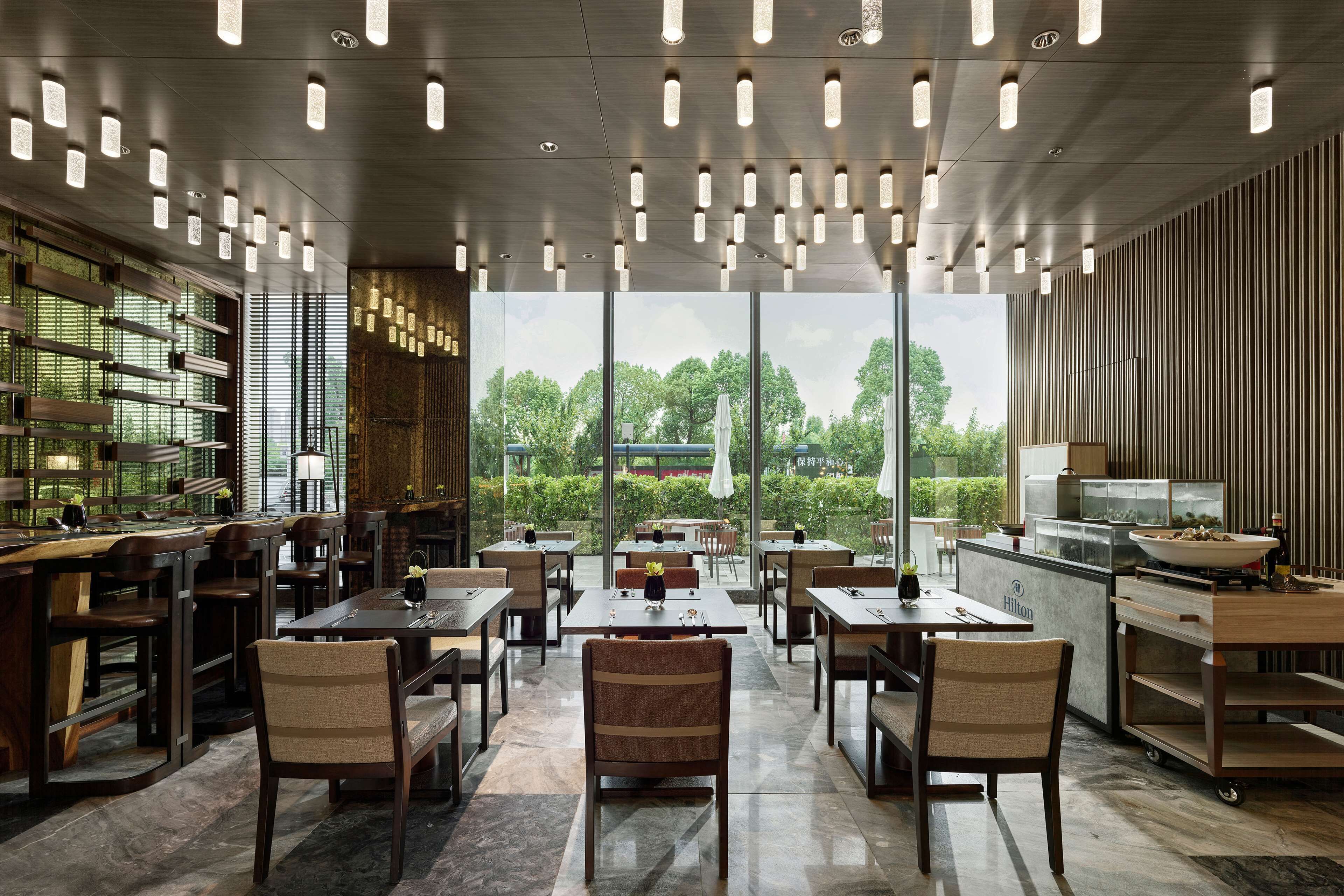A modern dining area with multiple tables and chairs, a bar area, and a view of greenery outside.
