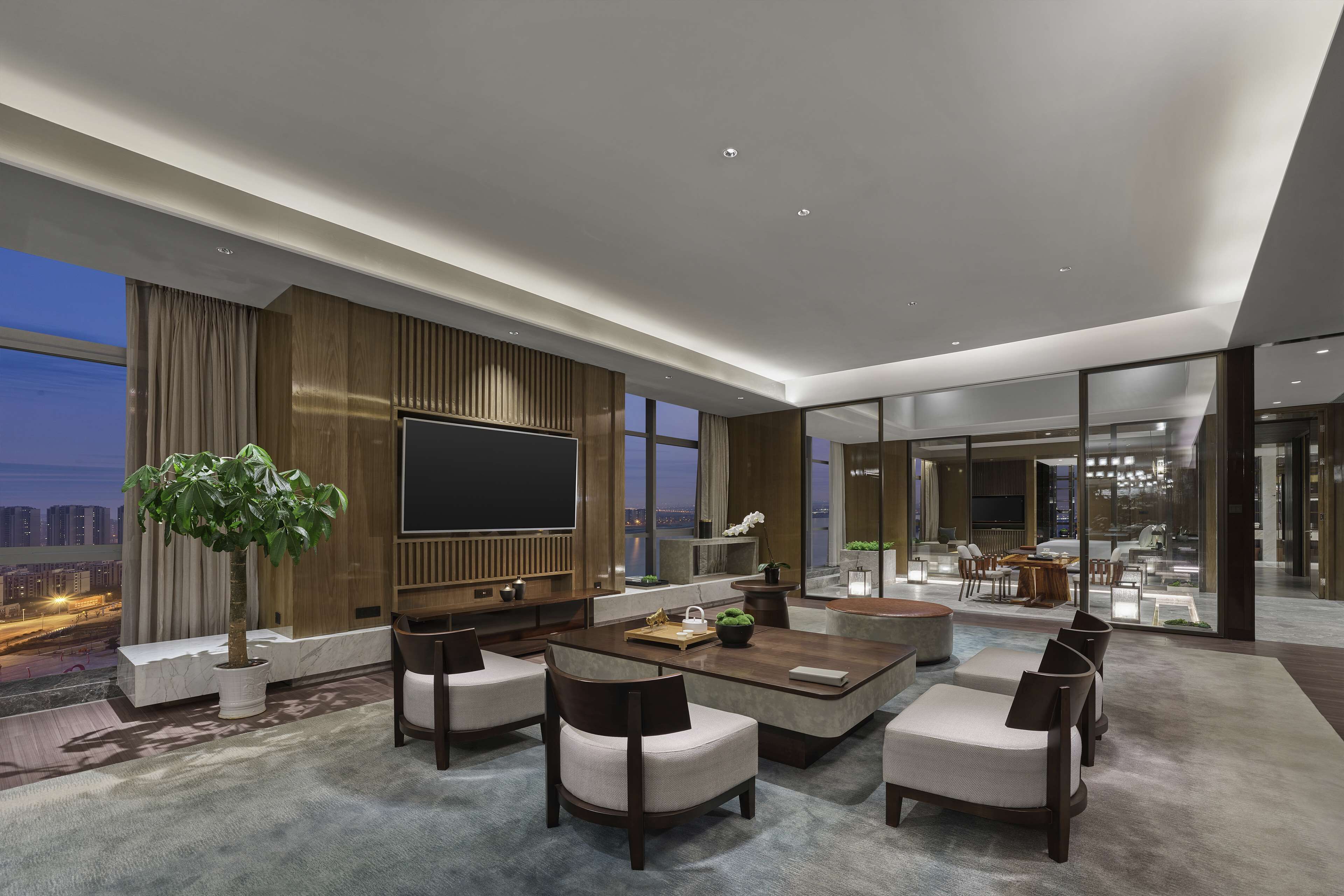 A modern living room with a large flat-screen TV, comfortable seating, a potted plant, and a view of the cityscape.