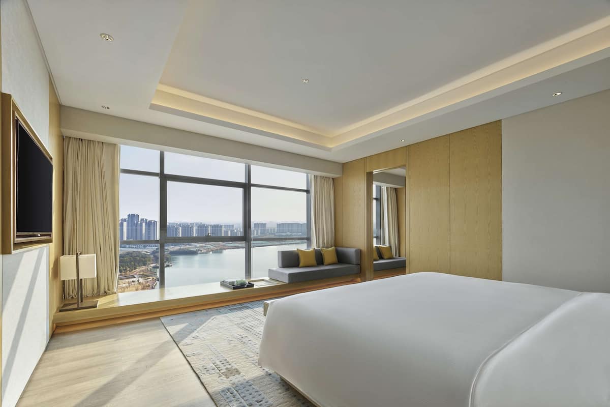 Une chambre d’hôtel moderne dotée d’un grand lit, d’un canapé, d’une télévision et offrant une vue sur le paysage urbain grâce à de grandes fenêtres.