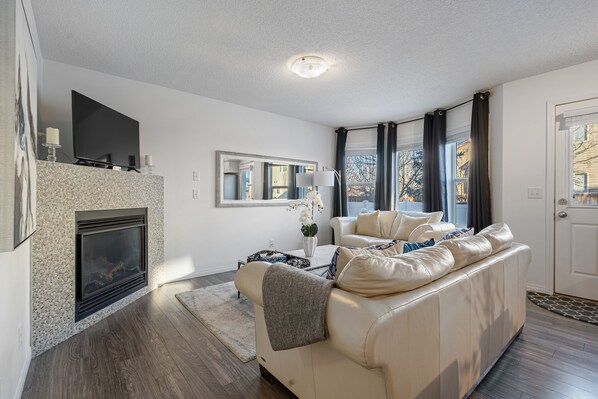 A modern living room with a fireplace, a sofa, a coffee table, and a large window with curtains. - Evergreen Calgary Townhome — 3BR, Sleeps 6, Gas Fireplace, Free Parking (Calgary)