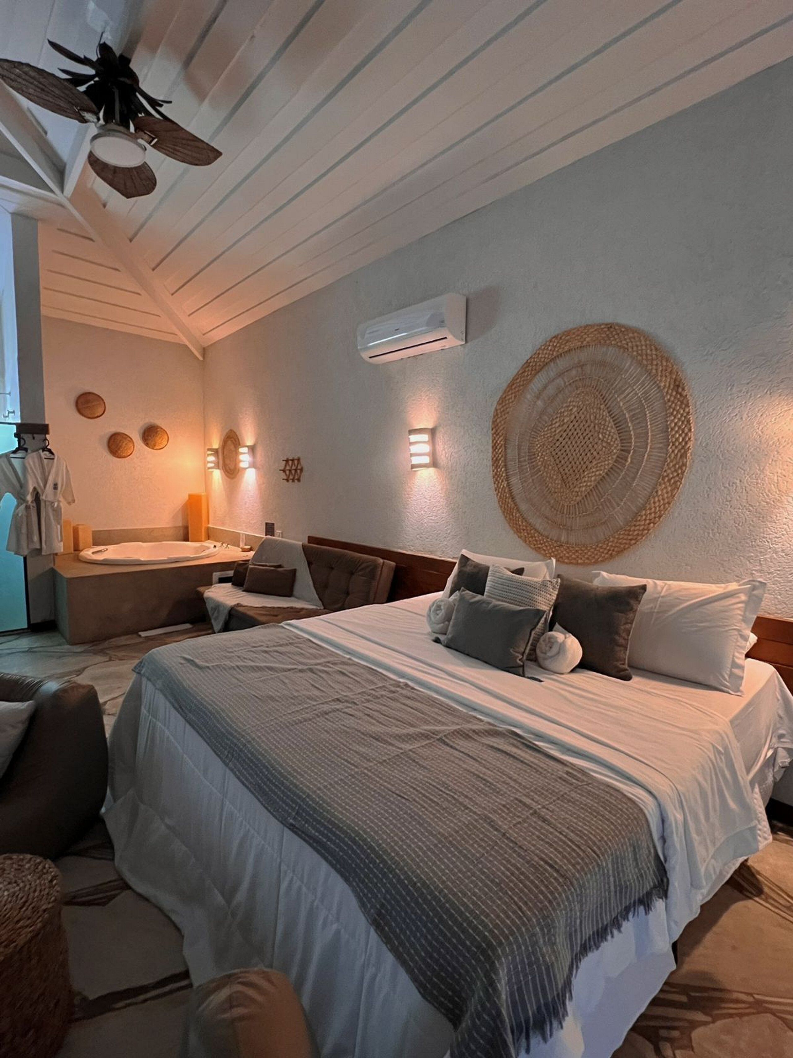 A bedroom with a large bed, a ceiling fan, a wall-mounted air conditioner, and a small seating area with a chair and a table.