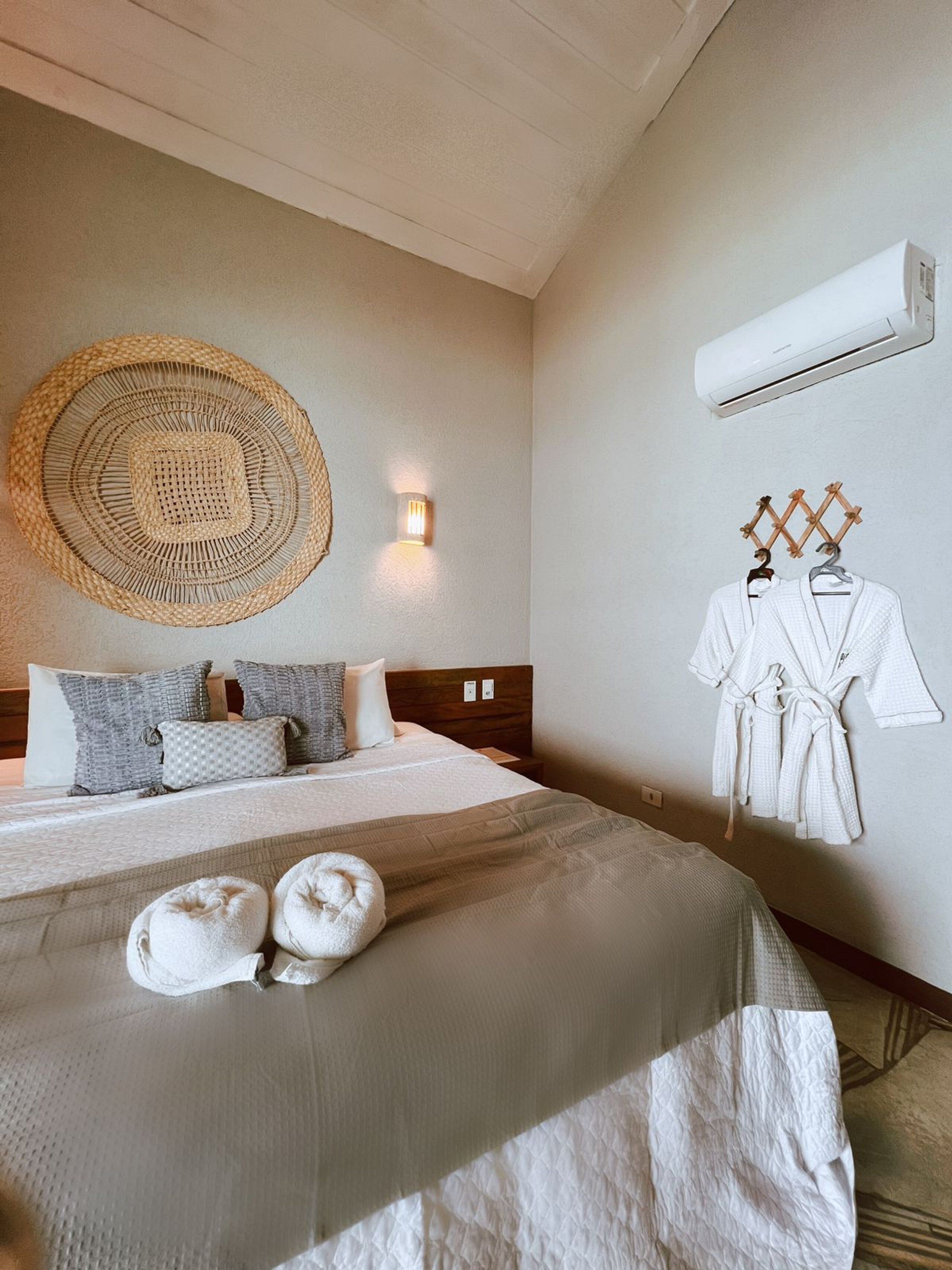 Una habitación de hotel con una cama, dos toallas, un aire acondicionado de pared y una lámpara mural, además de un tapiz de pared tejido.
