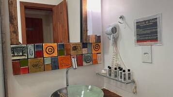 Uma casa de banho com um mosaico de azulejos coloridos, uma pia de cerâmica verde, uma toucador de madeira, um secador de cabelo e artigos de higiene numa estante.