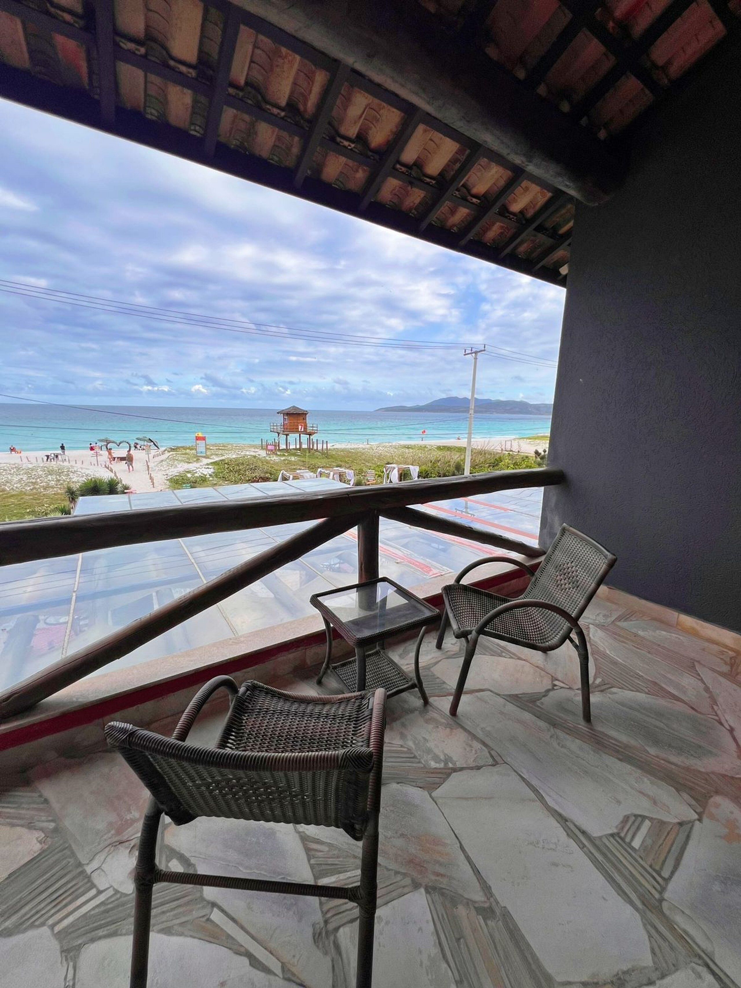 A balcony with wicker chairs and a glass table overlooking a beach with a lifeguard tower.