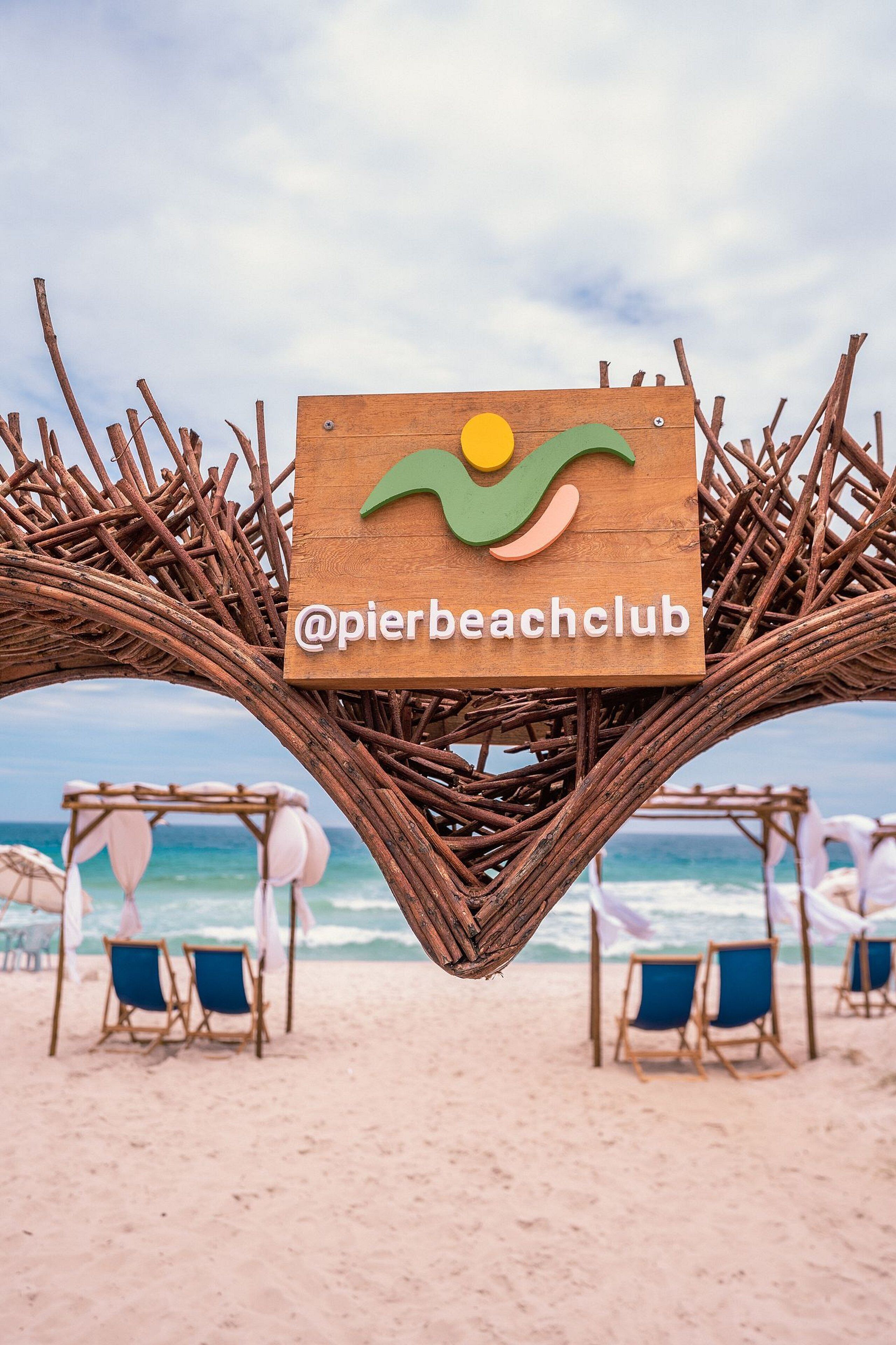 Rannalla oleva kyltti, jossa on logo ja teksti "@pierbeachclub", ja se on kiinnitetty puulevyyn. Kylttiä ympäröi punottu rakenne.