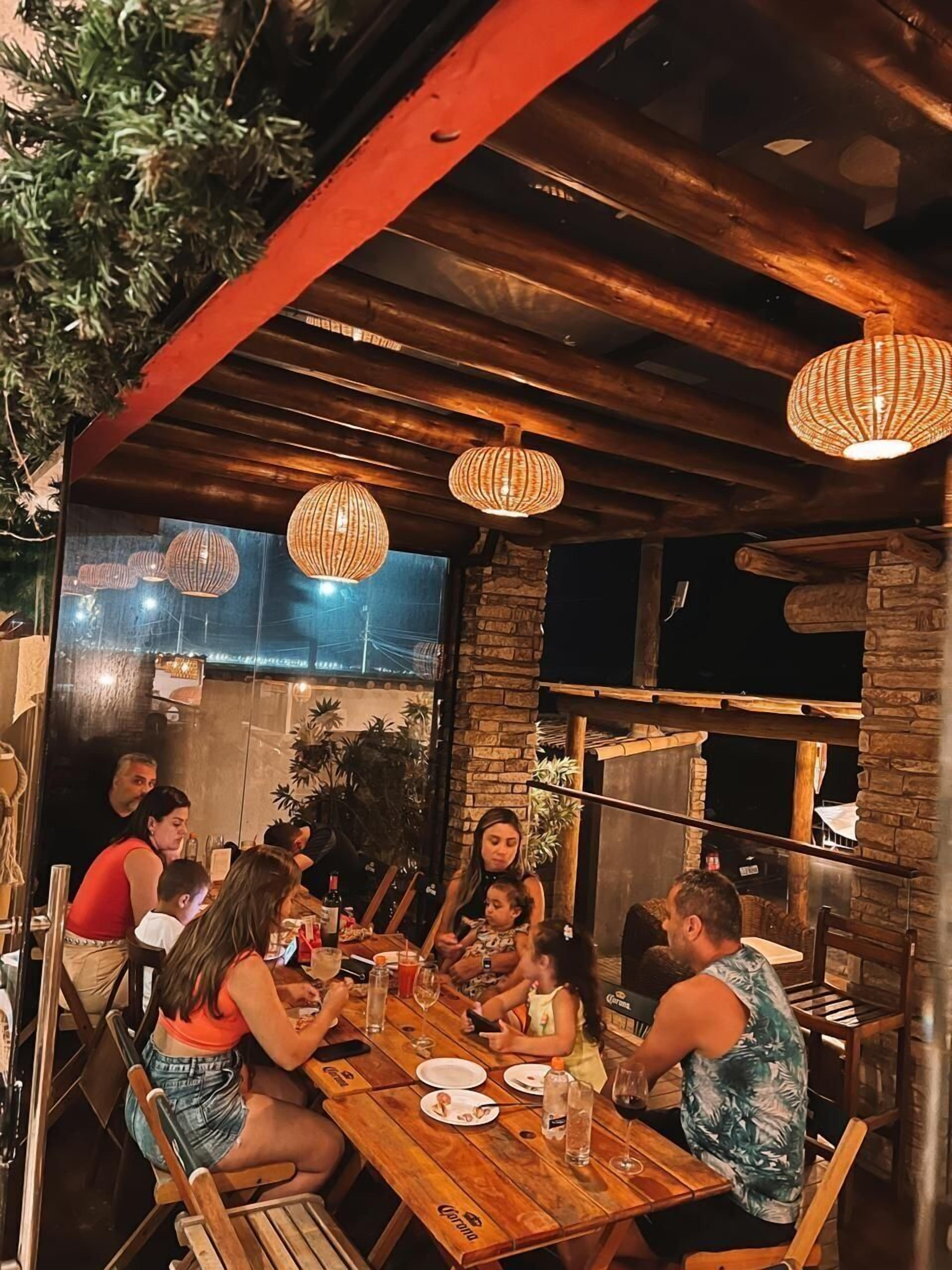 A group of people dining at an outdoor wooden table with hanging wicker lamps.
