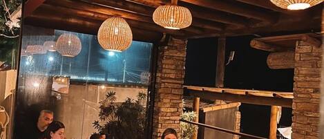 A group of people dining at an outdoor wooden table with hanging wicker lamps.