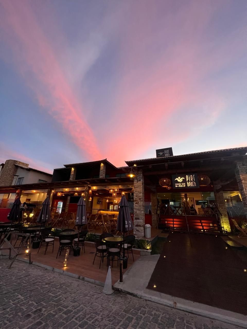 An outdoor seating area with tables and chairs, a stone building with a sign reading "Pier Beach", and a sunset sky.