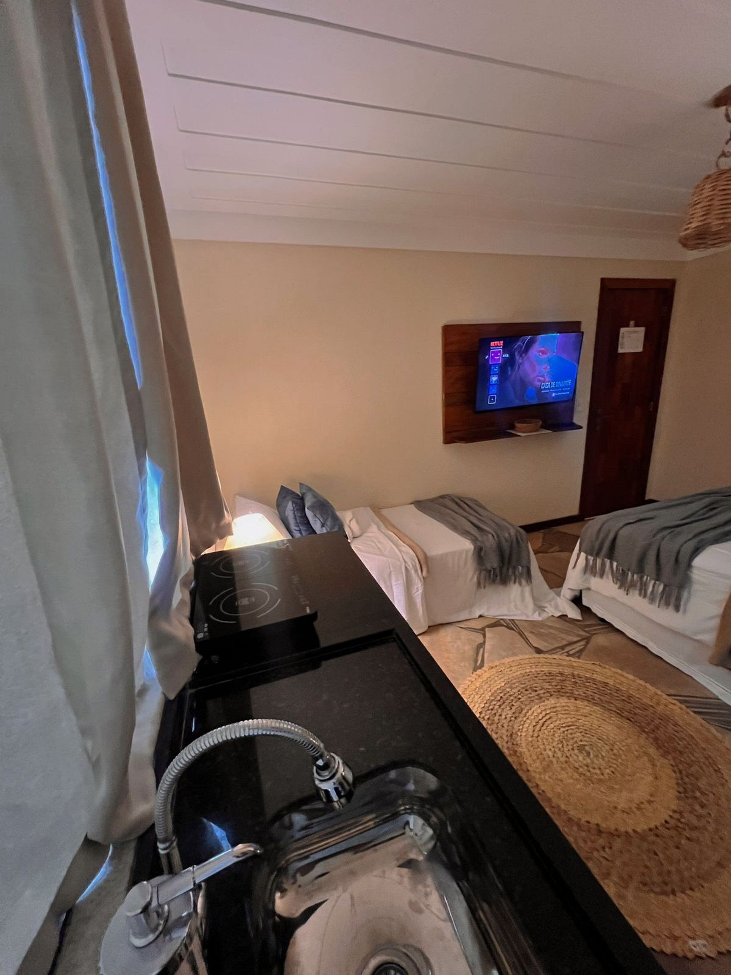 A hotel room with a kitchenette, a TV mounted on the wall, a bed with a blanket, and a woven mat on the floor.