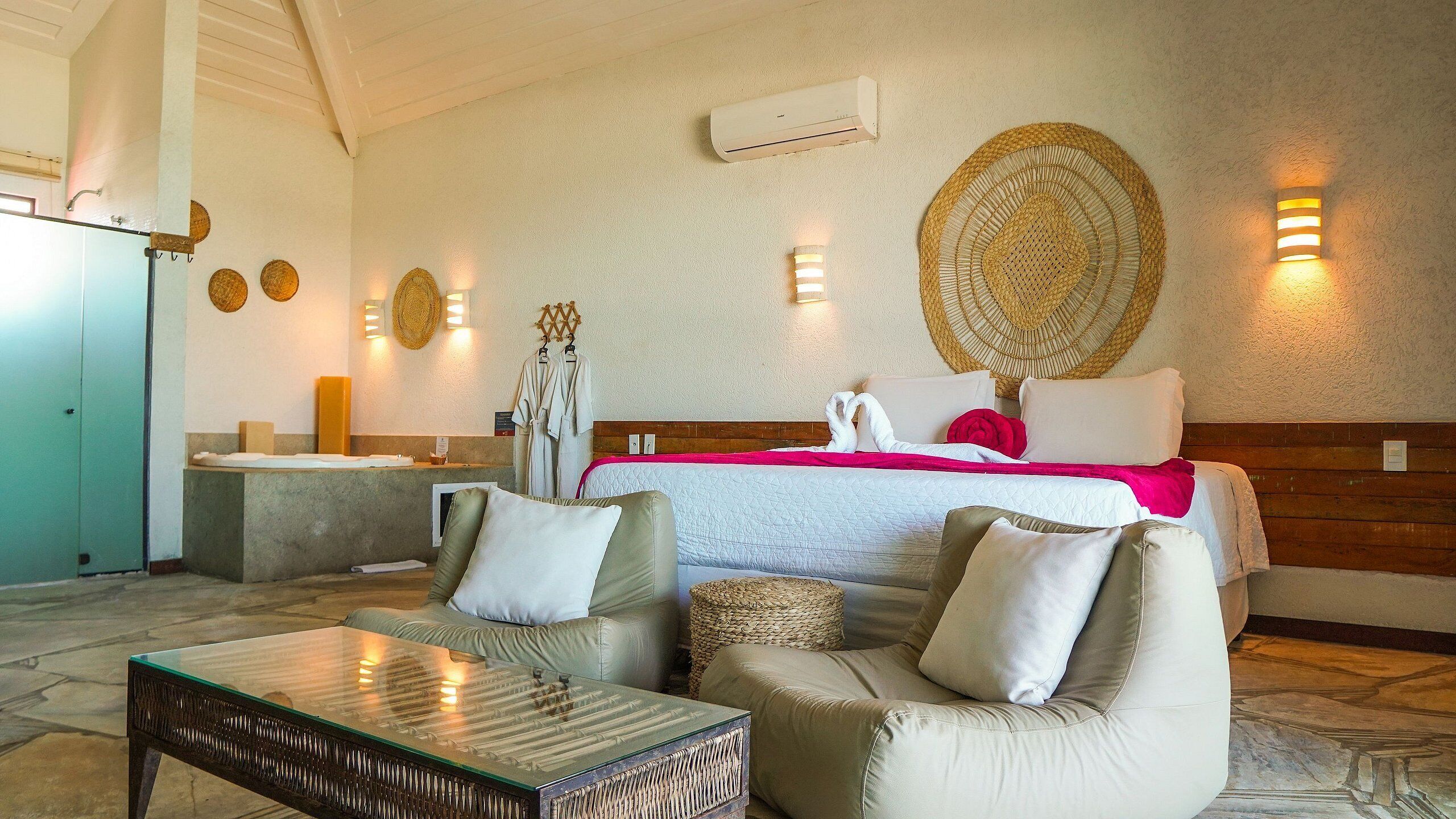 A hotel room with a bed, two armchairs, a glass coffee table, a small side table, a wall-mounted air conditioner, and a wall-mounted light fixture.