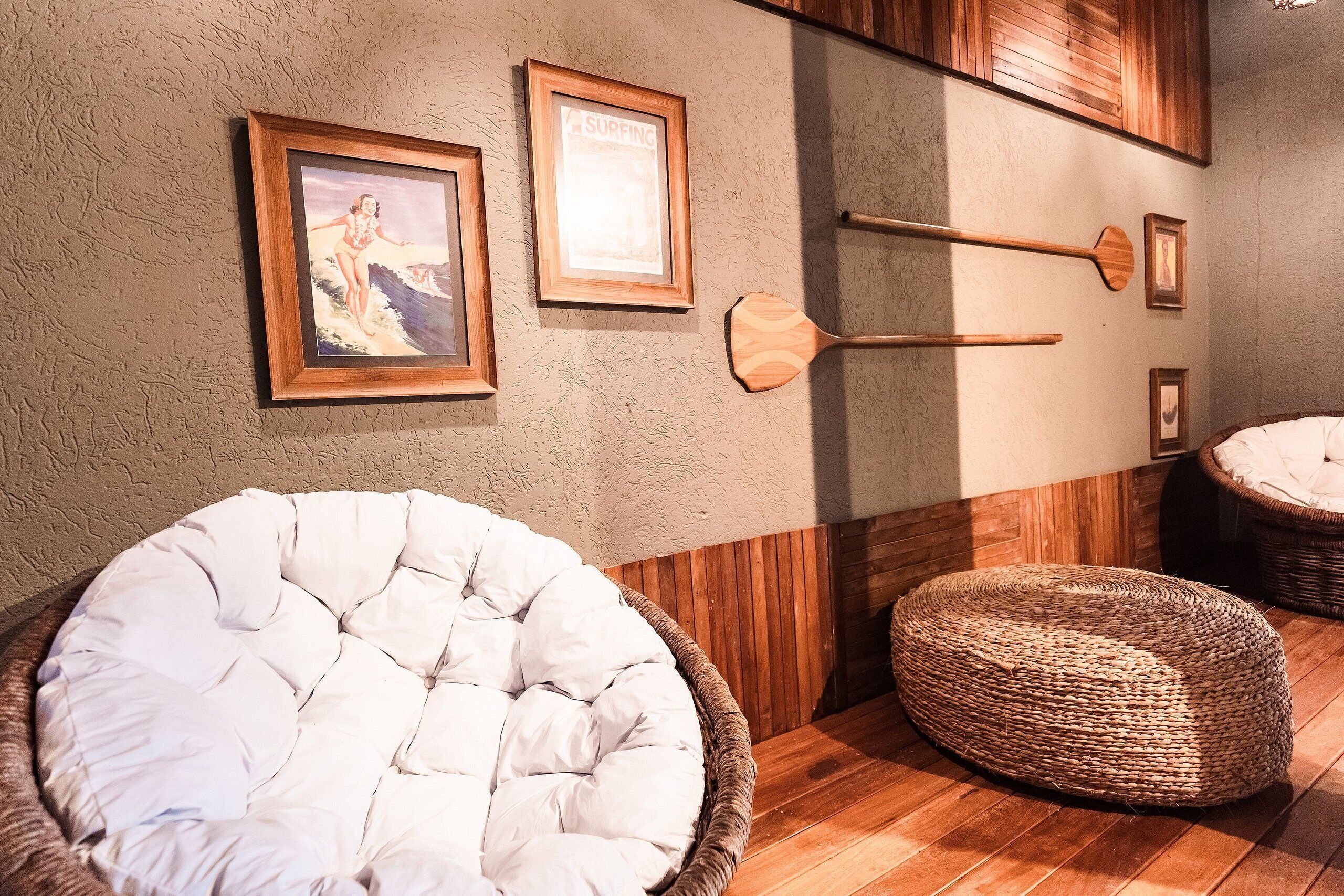 Een kamer met een houten vloer, een witte fauteuil en een rieten poef. Er hangen ingelijste foto's aan de muur en zijn houten wandlampen bevestigd.
