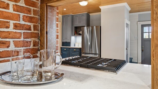 A kitchen with a brick wall, grey cabinets, a stainless steel refrigerator, and a tray with glasses.