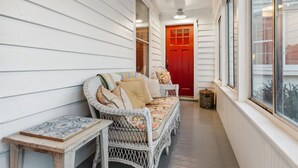 A narrow corridor with white wooden walls, a wicker sofa with cushions, a small table, and a red door at the end.
